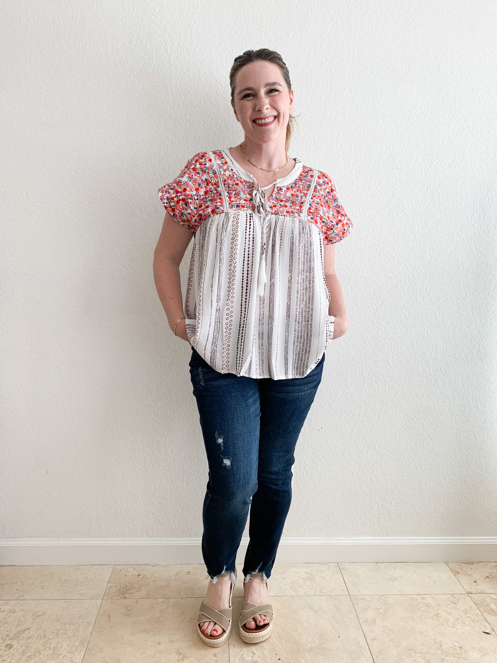 Smiling woman in front of white wall wearing flowy printed top and jeans.